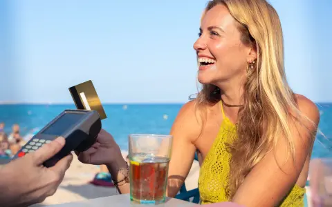 Lachende Frau bezahlt am Strand mit Karte. Selbst mit Krankengeld ist Urlaubsgeld drin!