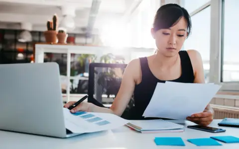 Junge Frau prüft Dokumente, vielleicht die Urlaubsbescheinigung des letzten Arbeitgebers, am Schreibtisch mit Laptop.
