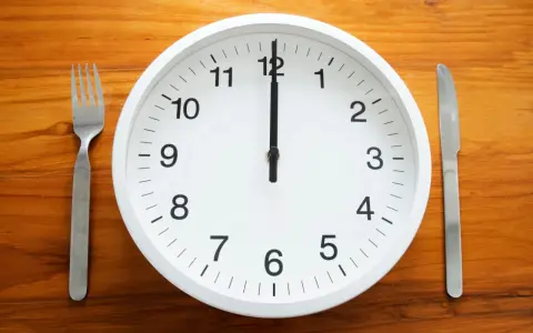 Uhr zeigt 12 Uhr. Gabel und Messer liegen bereit. Zeit für die Mittagspause während der Arbeitszeit.