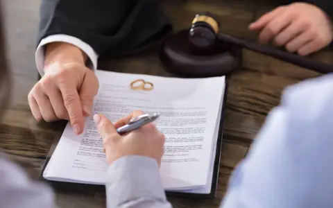 Gerichtlicher Hammer und Eheringe auf Scheidungsdokumenten. Ein Anwalt zeigt auf einen Paragraphen, während eine Person unterschreibt. Ein wichtiger Termin, der vielleicht Sonderurlaub erfordert.
