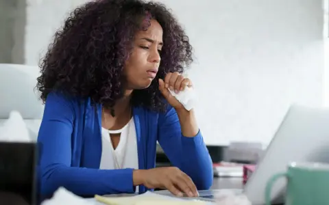 Frau hustet ins Taschentuch, während sie am Laptop arbeitet. Sie fragt sich, wie die Lohnfortzahlung bei einem Minijob auf Stundenbasis geregelt ist.