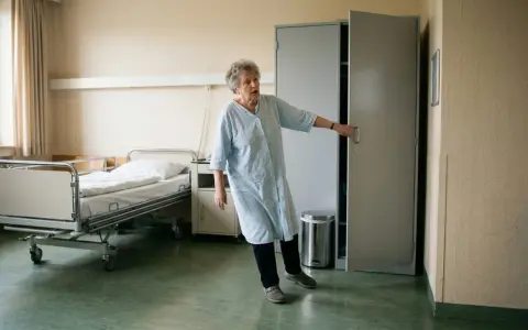Ältere Frau im Krankenhaushemd, die sich an einer Tür festhält. Sie hatte einen Arbeitsunfall, ging aber nicht zum Arzt.