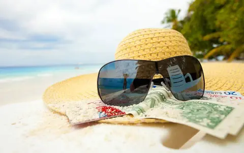 Sonnenbrille, Hut und Geld am Strand. Der Spiegel der Brille zeigt eine Person, die den Urlaub genießt. Ein Symbol für den **Urlaubsgeldanspruch**.