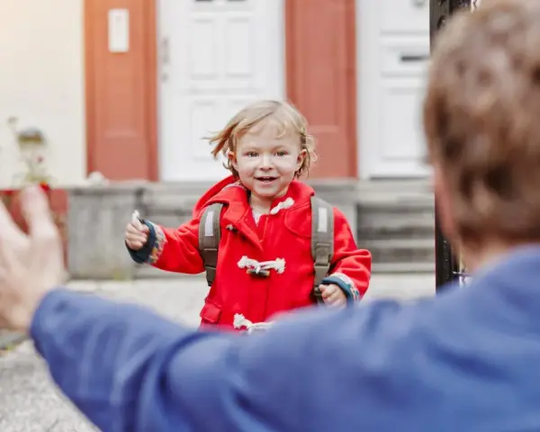 Kleines Mädchen im roten Mantel mit Rucksack rennt auf jemanden zu. Ein schönes Beispiel für das Wechselmodell bei Kindern.