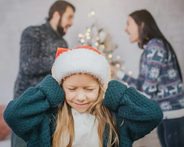 Ein Kind mit Weihnachtsmütze hält sich die Ohren zu, während die Eltern im Hintergrund streiten. Weihnachten trennen sie.
