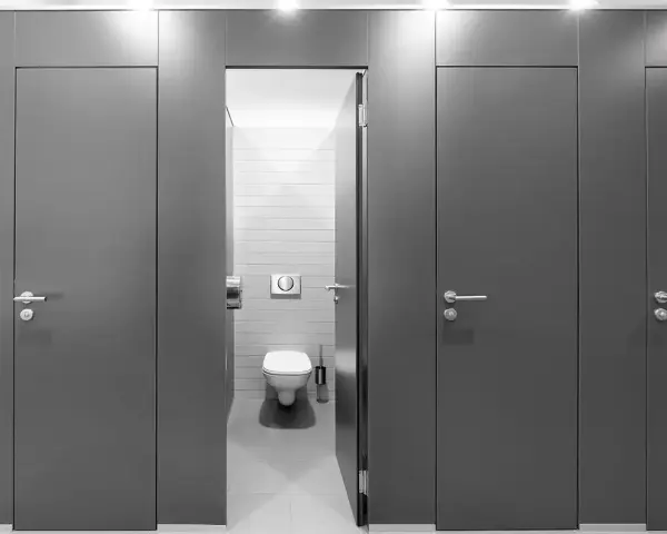 Reihe von grauen Türen zu Toilettenkabinen. Eine Tür ist geöffnet und gibt den Blick auf eine Toilette frei. Gute Frage, wie viele Toiletten pro Mitarbeiter?