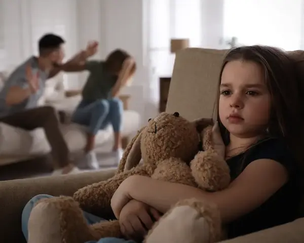 Ein trauriges Mädchen hält seinen Teddybär, während im Hintergrund Eltern streiten. Eine blitzscheidung ohne trennungsjahr hinterlässt Spuren.