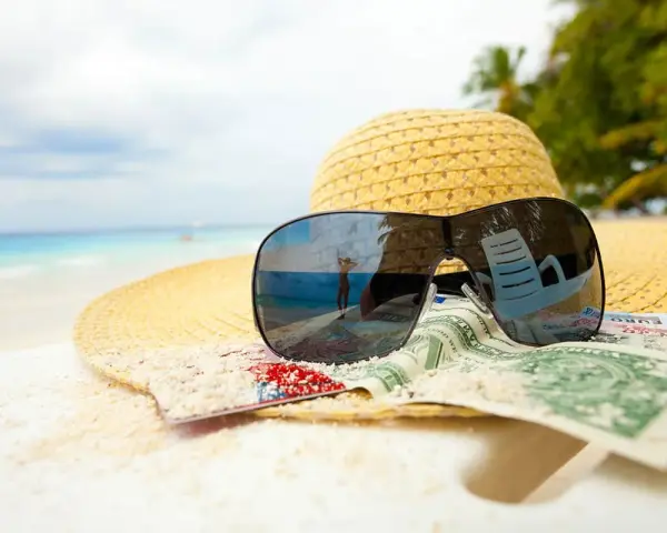 Sonnenbrille, Hut und Geld am Strand. Der Spiegel der Brille zeigt eine Person, die den Urlaub genießt. Ein Symbol für den **Urlaubsgeldanspruch**.