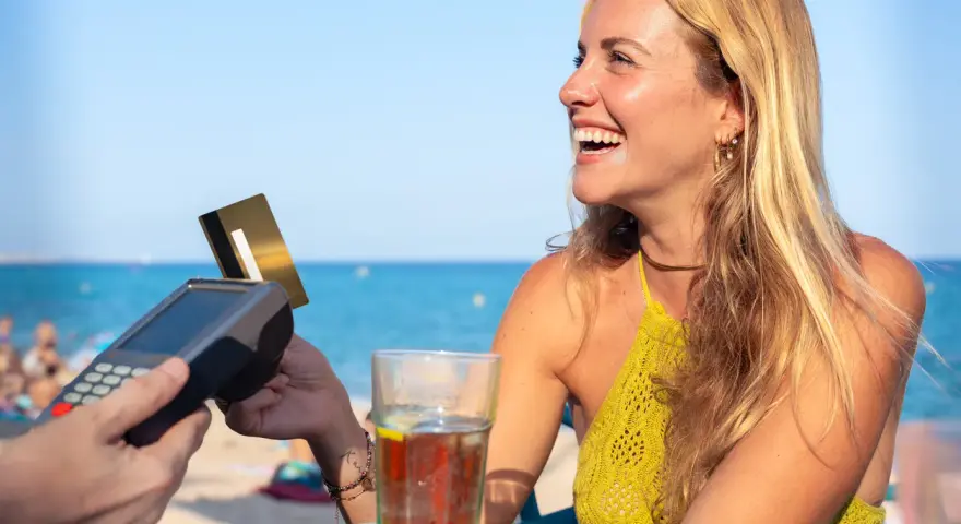 Lachende Frau bezahlt am Strand mit Karte. Selbst mit Krankengeld ist Urlaubsgeld drin!
