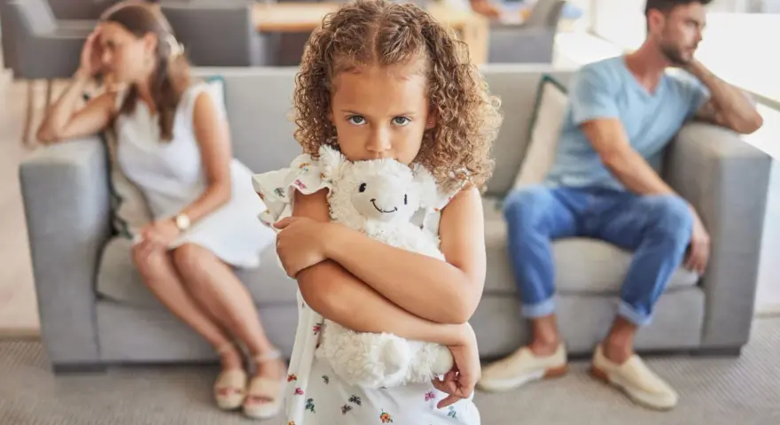 Ein Kind umklammert sein Stofftier, während die Eltern im Hintergrund getrennt sitzen. Ein Bild, das die emotionale Belastung eines Trennungsjahres mit Kindern widerspiegelt.