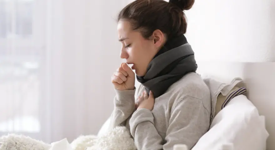Junge Frau hustet im Bett, eingewickelt in eine Decke. Sie hat einen Schal um den Hals. Die Szene könnte eine **Abmahnung wegen Krankheit** symbolisieren, wenn sie ihre Arbeit vernachlässigt.