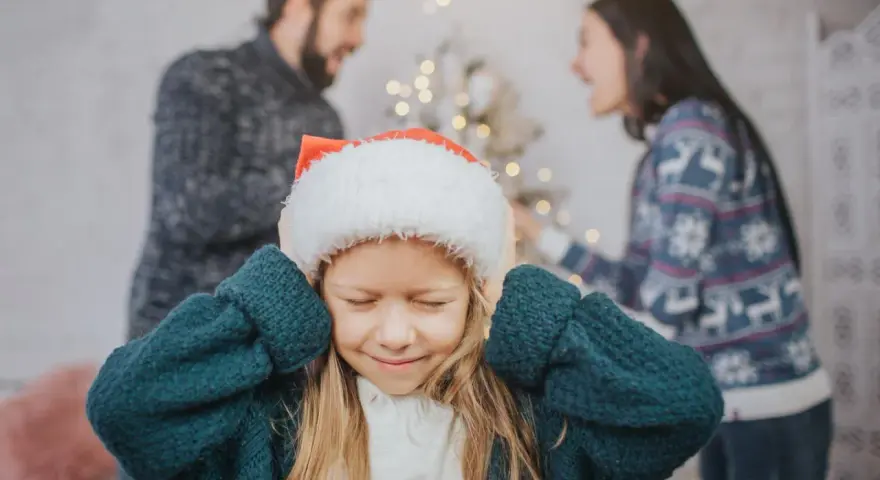 Ein Kind mit Weihnachtsmütze hält sich die Ohren zu, während die Eltern im Hintergrund streiten. Weihnachten trennen sie.