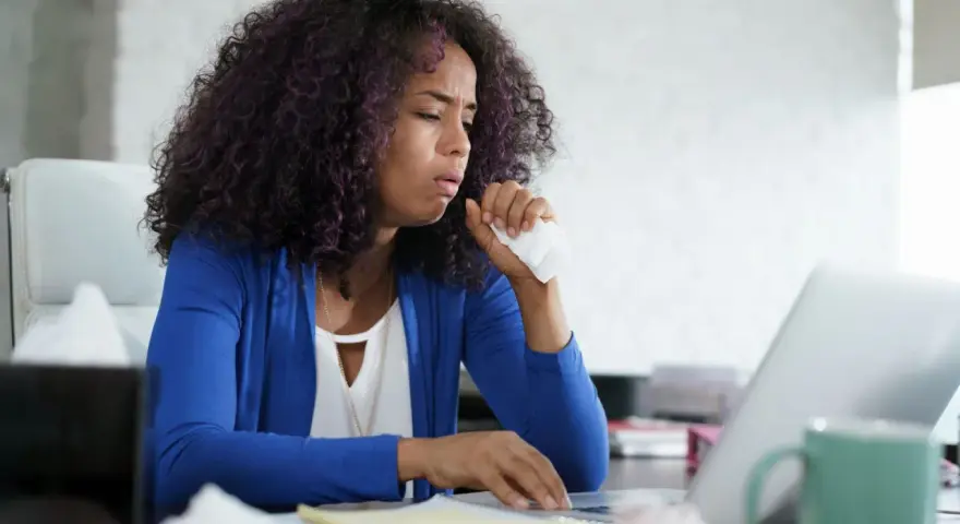 Frau hustet ins Taschentuch, während sie am Laptop arbeitet. Sie fragt sich, wie die Lohnfortzahlung bei einem Minijob auf Stundenbasis geregelt ist.