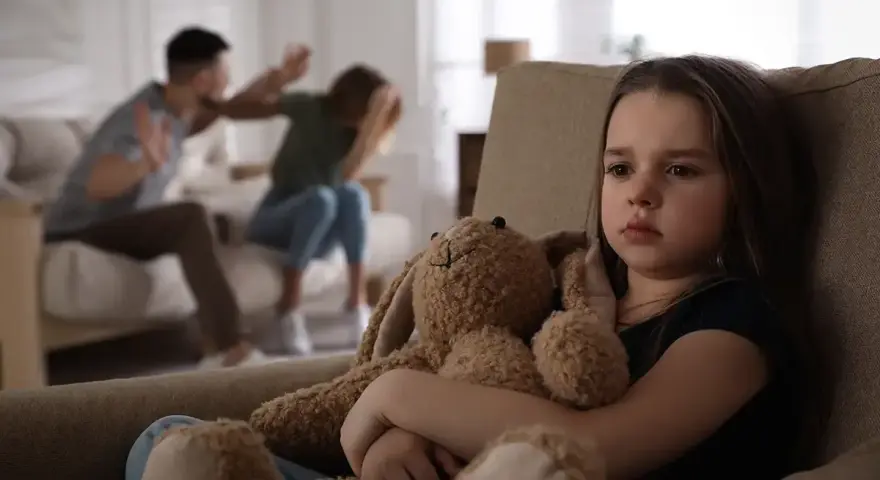 Ein trauriges Mädchen hält seinen Teddybär, während im Hintergrund Eltern streiten. Eine blitzscheidung ohne trennungsjahr hinterlässt Spuren.