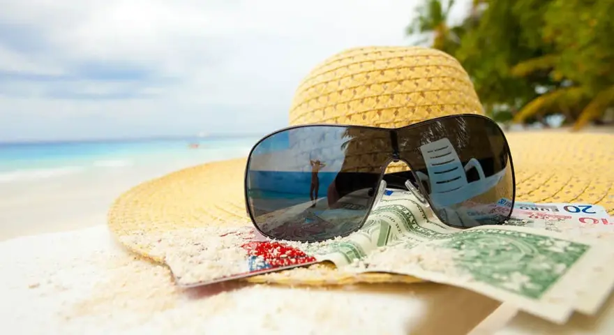 Sonnenbrille, Hut und Geld am Strand. Der Spiegel der Brille zeigt eine Person, die den Urlaub genießt. Ein Symbol für den **Urlaubsgeldanspruch**.