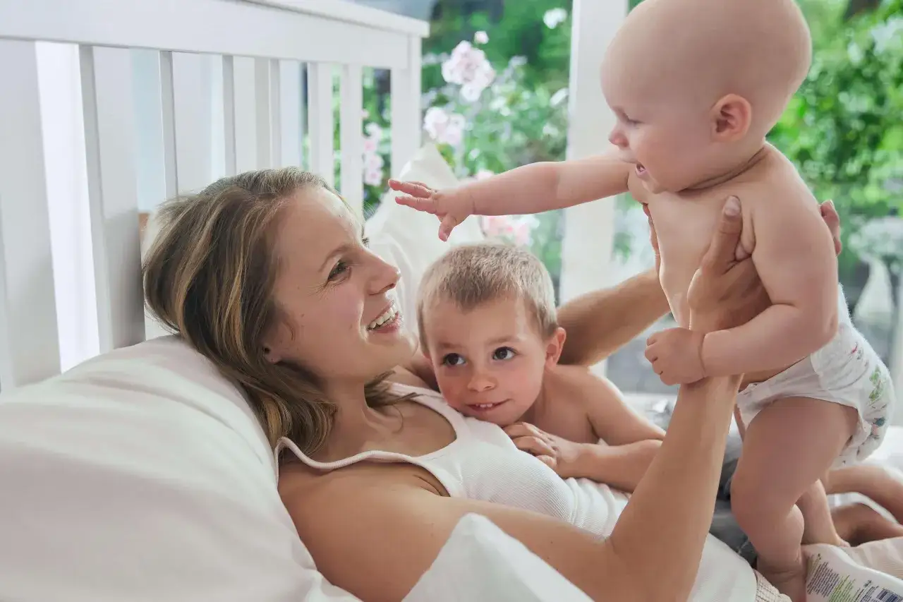 Glückliche Familie im Bett. Ein Vater hält sein Baby hoch, während die Mutter und das ältere Kind zuschauen. Ein schönes Bild für den Vaterschaftsurlaub in Deutschland.