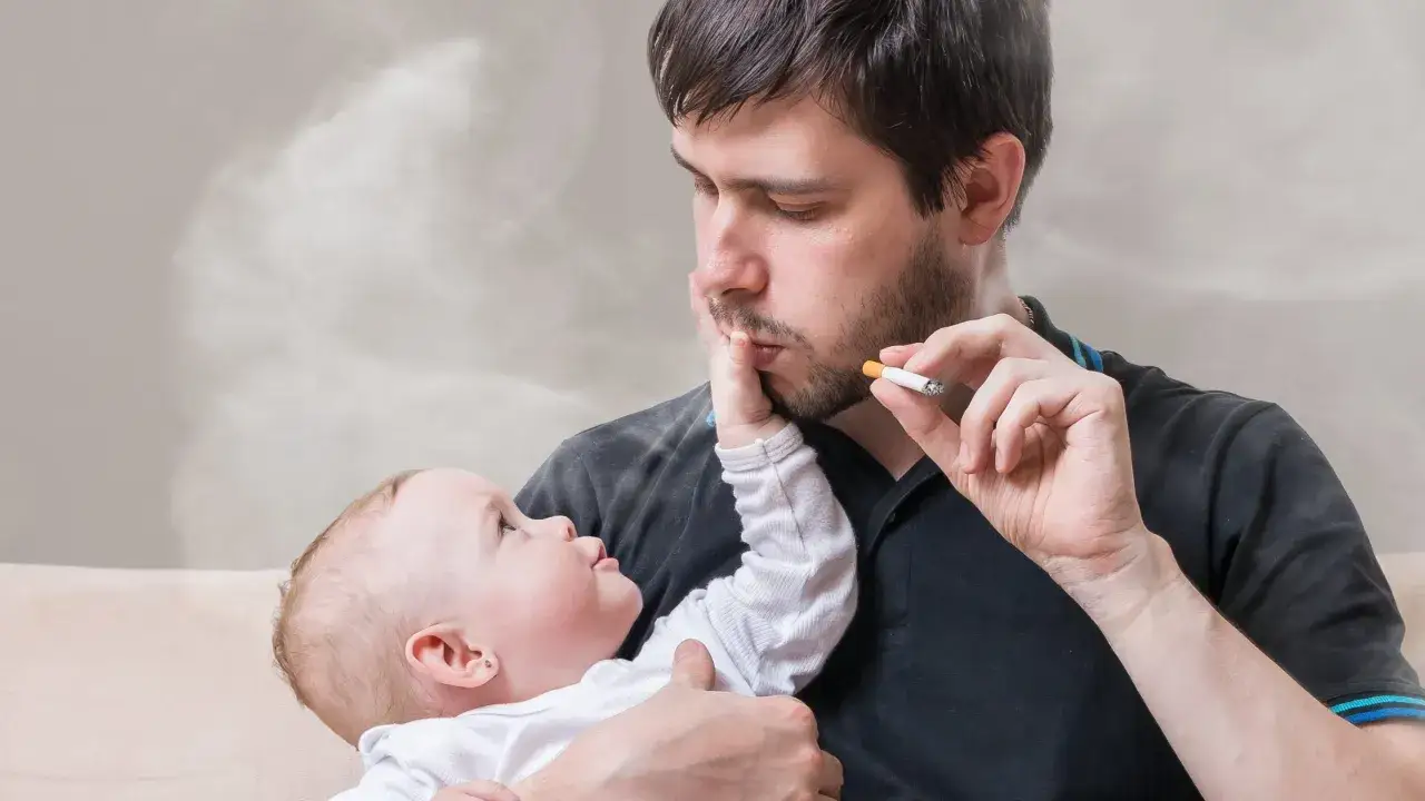 Mann hält Baby und raucht in der Wohnung. Rauch umgibt sie. Das Jugendamt könnte eingreifen.